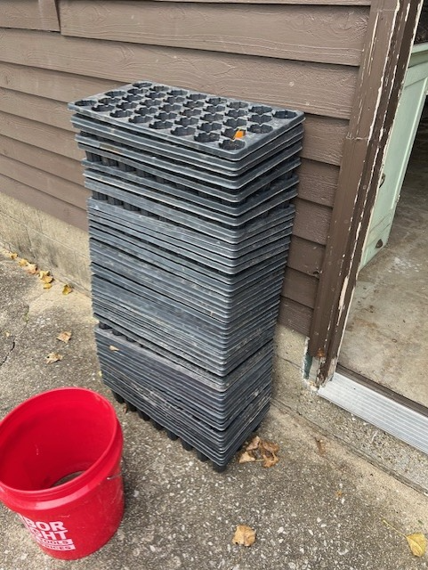 A stack of black, empty flats and a red 5-gallon bucket next to them 
